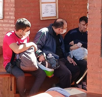 Ismael Obregón Olmedo en chaqueta oscura, sentado entre dos hombres con mascarillas, en un lugar de ladrillos al aire libre.