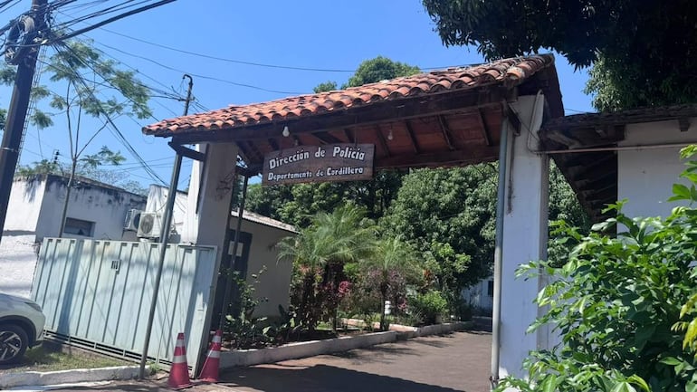 Entrada de la Dirección de Policía, edificio de techo a dos aguas rodeado de naturaleza, con un vehículo aparcado a la izquierda.