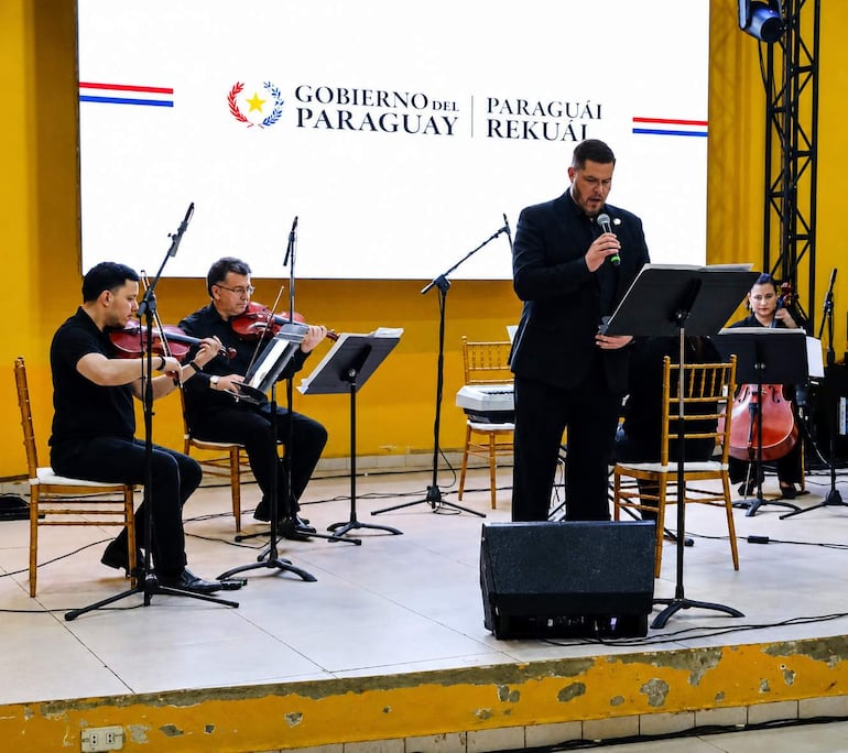 Hombre en traje oscuro habla con micrófono, flanqueado por violinistas y una chelista en un escenario decorado.