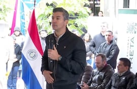 Santiago Peña Palacios en chaqueta negra, sostiene un micrófono mientras habla a la audiencia, con bandera de Paraguay de fondo.