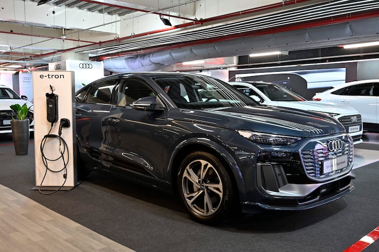 Modelo Audi e-tron en exhibición junto a estación de carga, con fondo neutro y otros vehículos Audi visibles.