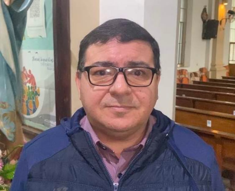 Padre Sergio Maciel con chaqueta azul y gafas, de pie en el interior de una iglesia con bancos visibles.