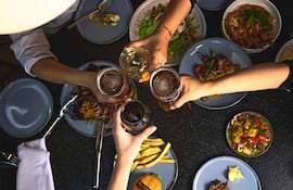 Cinco manos levantan vasos con cerveza y vino en una mesa llena de comida en un ambiente festivo.