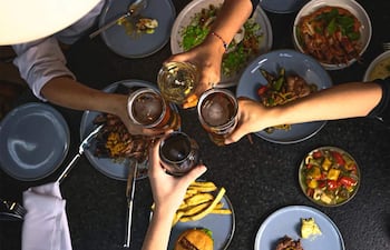 Cinco manos levantan vasos con cerveza y vino en una mesa llena de comida en un ambiente festivo.