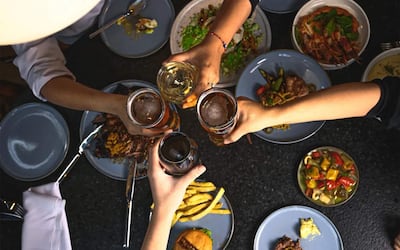 Cinco manos levantan vasos con cerveza y vino en una mesa llena de comida en un ambiente festivo.