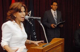 Myriam Peña, con blusa blanca y micrófono, gesticula mientras un hombre en traje gris la observa en un auditorio.
