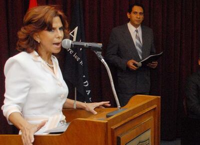 Myriam Peña, con blusa blanca y micrófono, gesticula mientras un hombre en traje gris la observa en un auditorio.