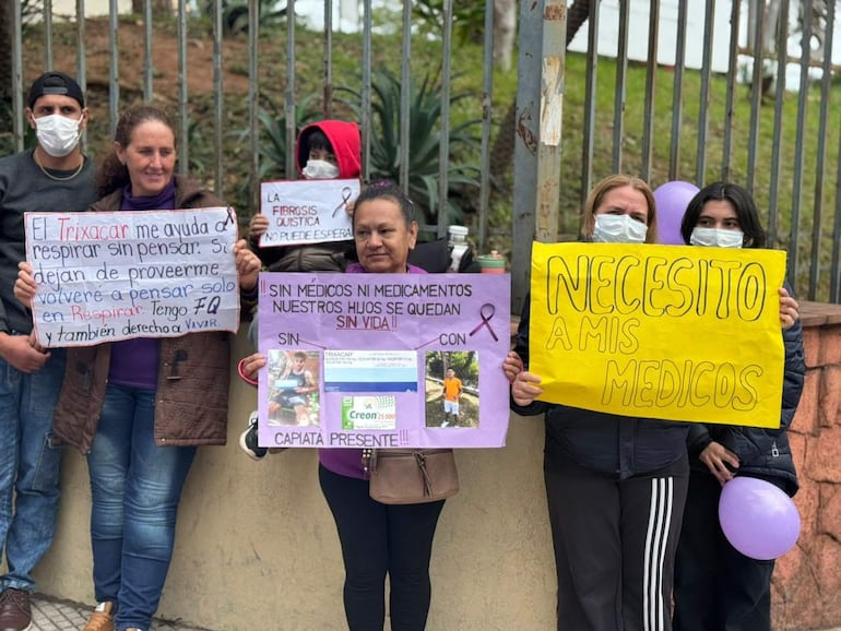 Grupo de cinco personas con mascarillas, sosteniendo carteles sobre fibrosis quística y necesidades médicas, en un ambiente de manifestación pacífica.