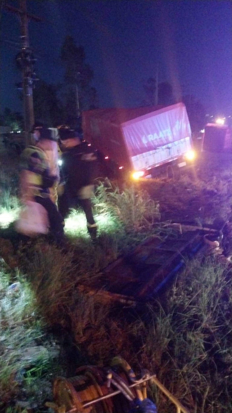 Dos miembros de emergencia en atuendo de protección en la escena de un accidente con un camión volcado de color rojo y hierba alrededor.