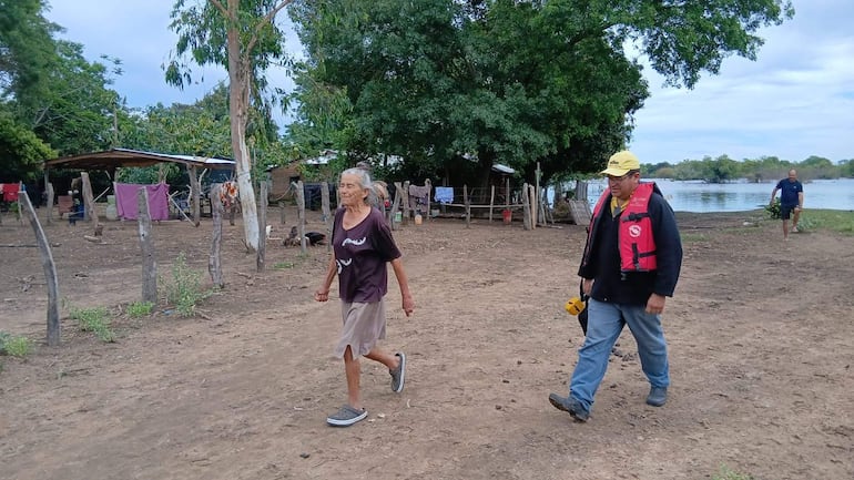 Mujer canosa con camiseta oscura y falda clara sostiene la mano de un hombre con chaleco salvavidas y gorra amarilla, caminando en entorno rural.