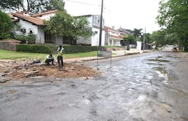Dos trabajadores con chalecos de seguridad, uno usando pala, el otro en el suelo. Escena de trabajo en una calle con casas y vegetación de fondo.