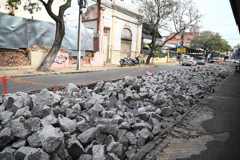 Rocas grises apiladas en la calle, conos naranjas delimitan área de obra. Dos motocicletas estacionadas a la izquierda.