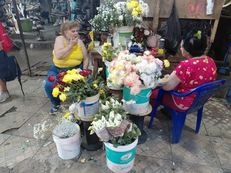 Dos mujeres en un mercado de flores, una con blusa amarilla y otra con blusa roja, rodeadas de arreglos florales coloridos.