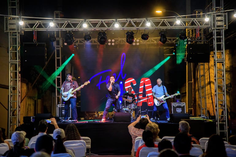 Cantante dinámico con micrófono en mano, dos guitarristas y un baterista, luces del escenario en un ambiente festivo.