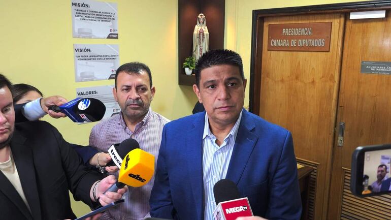 Hugo Meza en blazer azul habla mientras Miguel Del Puerto escucha atentamente, rodeados de periodistas en un entorno formal.