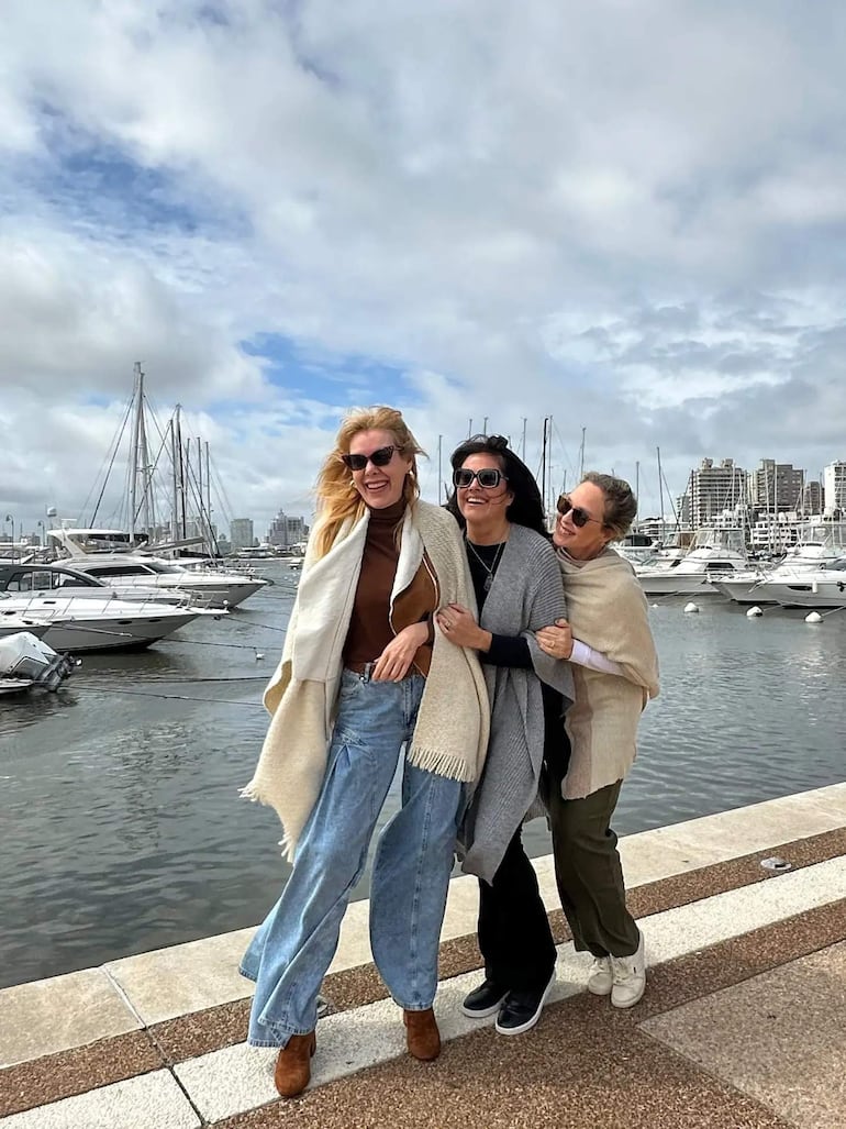 Mujer rubia con gafas de sol y prenda clara sonríe junto a dos amigas en un puerto con yates de fondo.