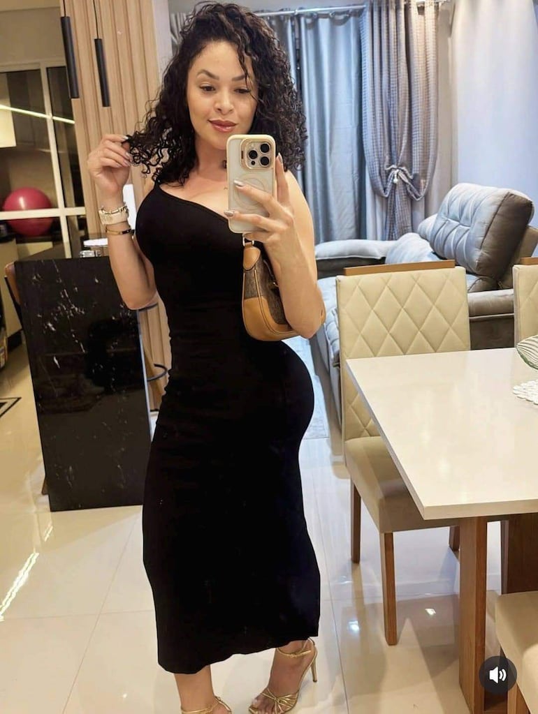 Mujer con cabello rizado y vestido negro ajustado, tomando una autofoto en un ambiente moderno con muebles claros y decorativos.