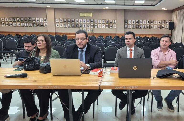 Mujer en blusa verde y hombre en traje oscuro conversan en mesa, mientras otros observan con laptops en sala vacía.