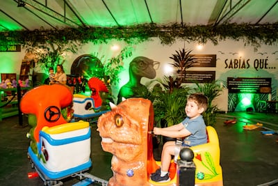 Niño con camiseta azul en carrito de dinosaurio, mientras mujer con bebé lo observa en fondo festivo con decoraciones vegetales.