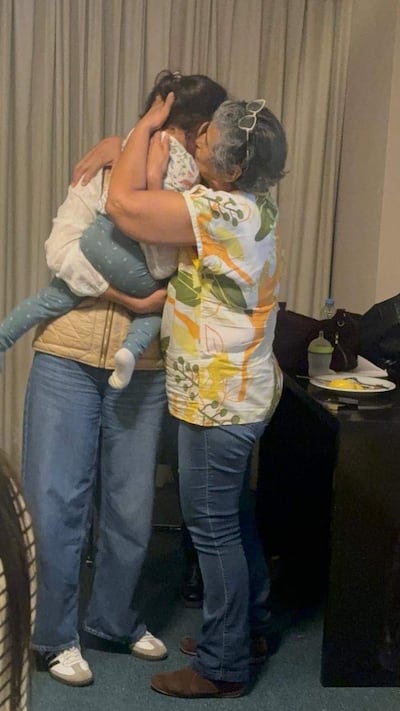 Dos mujeres en camiseta blanca y blusa clara, abrazándose con una niña entre ellas en un entorno hogareño.