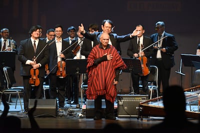 Músicos en el escenario, con hombre mayor en poncho resaltando respeto, mientras otro dirige con gestos entusiastas.