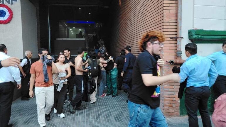 Varias personas en ropa casual se agrupan cerca de la entrada del edificio, mostrando preocupación mientras algunos se abrazan.