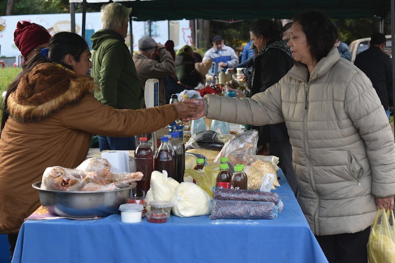 Mujer con abrigo marrón y bufanda estrecha mano a otra con abrigo gris claro en un mercado al aire libre.