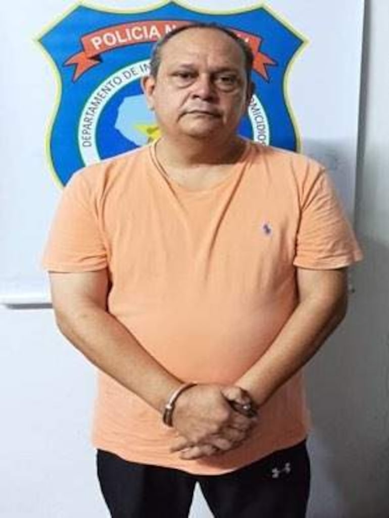 Hombre esposado con camiseta naranja en una comisaría, frente a un escudo policial, postura erguida y expresión tranquila.