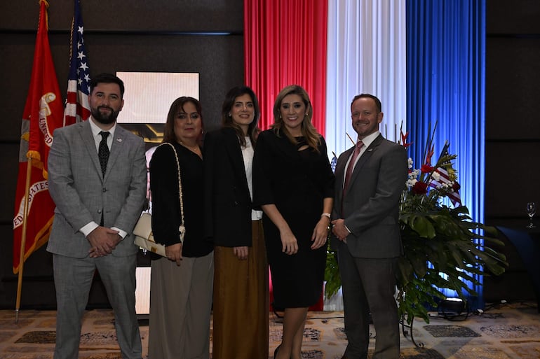 Grupo de cinco personas sonrientes, vestidas formalmente, posan en un salón decorado con banderas y plantas.