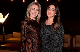 Dos mujeres sonrientes posan juntas, una en vestido de diseño marrón y negro, otra en vestido negro con brillo, en ambiente decorado con luces suaves.