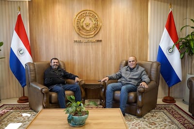 Basilio Núñez sonríe mientras conversa con Miguel Del Puerto en una oficina decorada con banderas paraguayas.