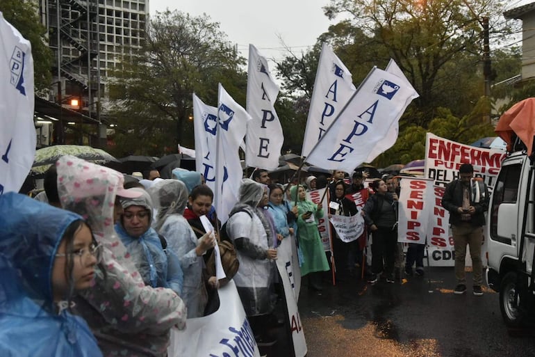 Grupo de manifestantes bajo la lluvia, con impermeables, sosteniendo pancartas y banderas frente al Ministerio de Salud.
