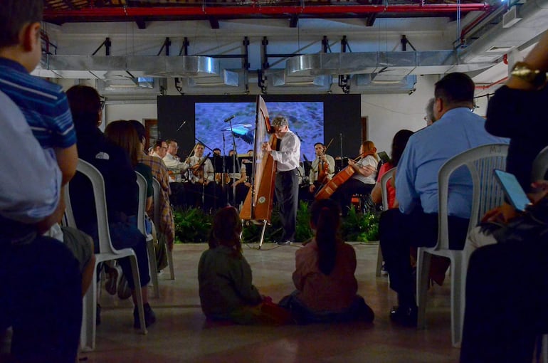 Músicos en el escenario, entre ellos un arpista con camisa blanca. Público atento, incluyendo niños con ropa de colores distintos.