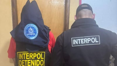 Dos personas visten chaquetas de Interpol; uno lleva capucha y chaleco con la inscripción 'INTERPOL DETENIDO'.