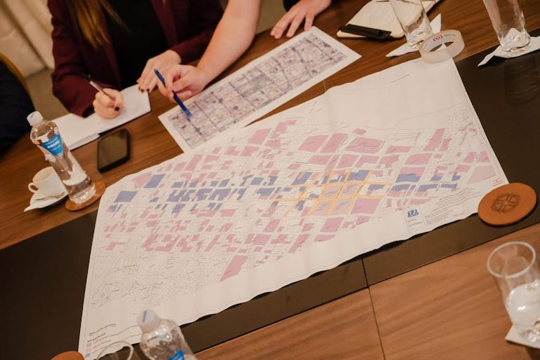 Reunión formal con dos personas, una con blusa negra y otra en prenda burdeos, analizando mapas y documentos en una mesa.