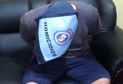 Sospechoso cubierto con paño azul y la palabra 'HOMICIDIOS', sentado en un sofá, vestido con camiseta gris y pantalones oscuros.