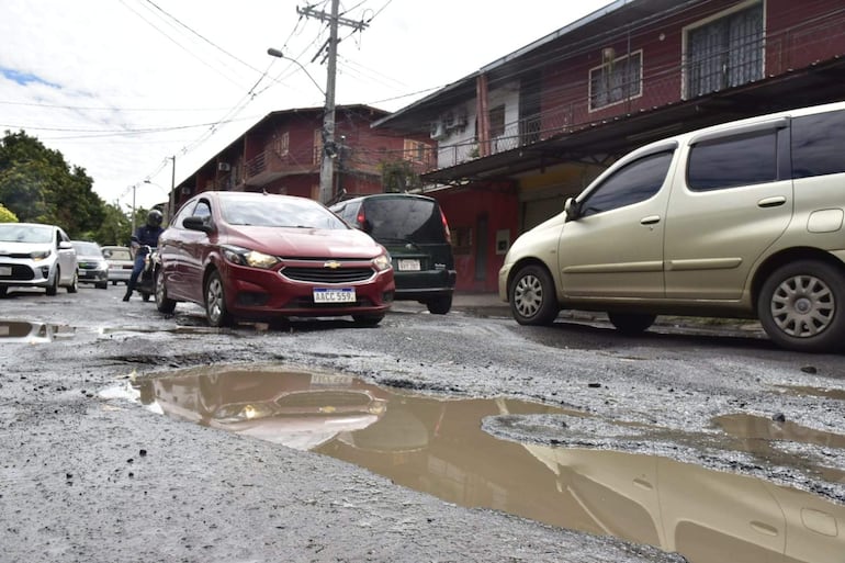 Un auto rojo y una camioneta verde transitan por una calle con baches y charcos en un ambiente urbano.