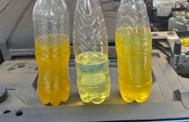 Tres botellas de plástico sobre un motor de automóvil, con líquidos amarillentos, en un entorno exterior despejado.