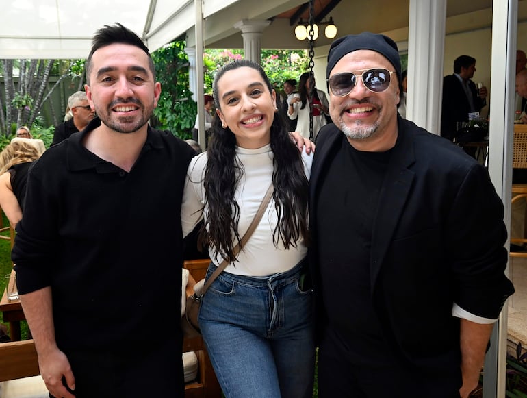 Hombre con barba y camisa negra, mujer con blusa blanca y jeans, y hombre con gorro negro y gafas, todos sonrientes en un entorno festivo.