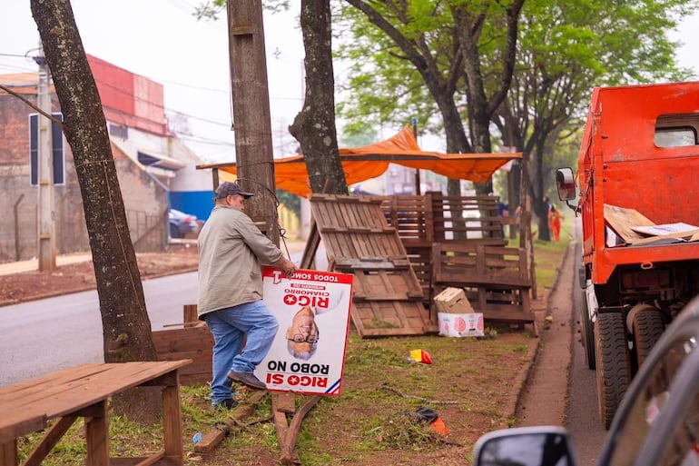 Hombre robusto en chaqueta y jeans sosteniendo un cartel al revés en una calle, con un camión rojo al fondo.