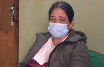 Mujer con mascarilla azul y chaqueta oscura, mostrando expresión seria en un ambiente doméstico con pared verde.