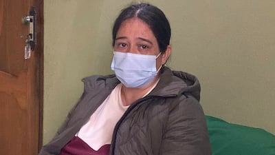 Mujer con mascarilla azul y chaqueta oscura, mostrando expresión seria en un ambiente doméstico con pared verde.