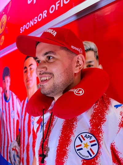 Hombre con camiseta de rayas rojas y blancas y gorra roja que dice 'ROJA', sonríe frente a imágenes de jugadores en un fondo gráfico rojo.