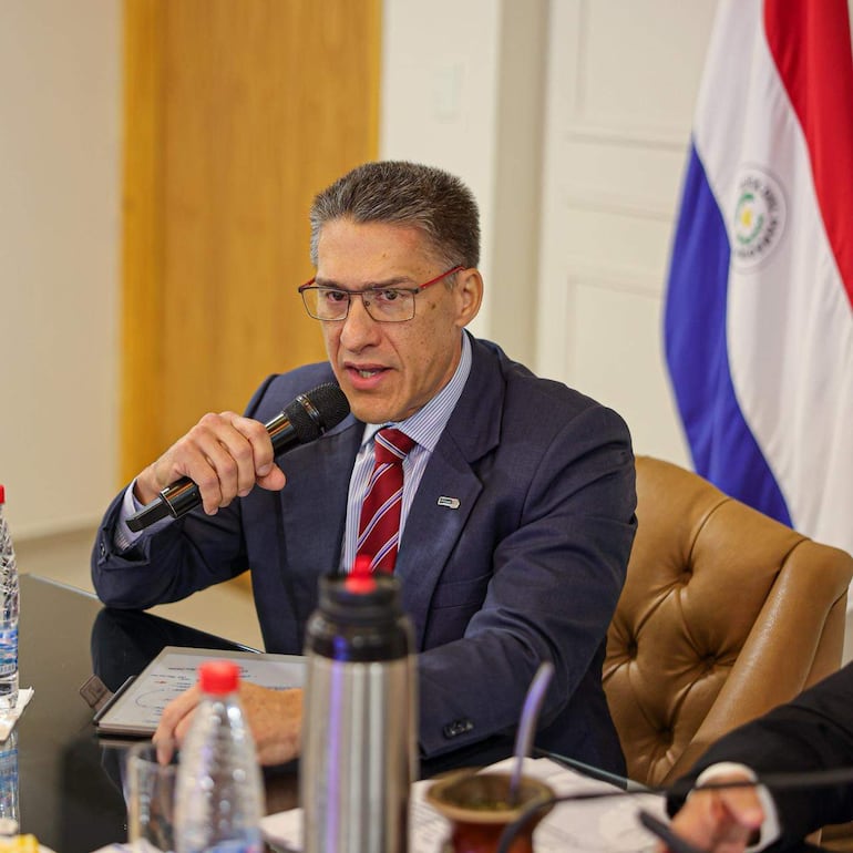 Basilio Núñez habla sosteniendo un micrófono, vestido con traje oscuro y corbata a rayas, en un ambiente formal con bandera paraguaya.
