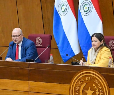 Óscar Lovera en traje azul y corbata roja habla frente a una mujer de abrigo amarillo, en recinto con banderas de Paraguay.