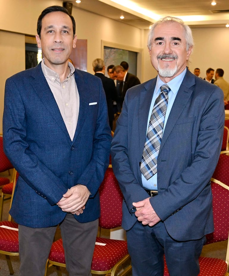Hombre en blazer azul oscuro y otro en traje gris, ambos sonriendo en un salón de eventos con sillas rojas.