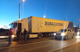 Cinco hombres, dos hablando con el conductor de un camión negro, todo en un entorno de carretera bajo un cielo crepuscular.