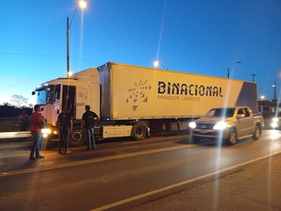 Cinco hombres, dos hablando con el conductor de un camión negro, todo en un entorno de carretera bajo un cielo crepuscular.