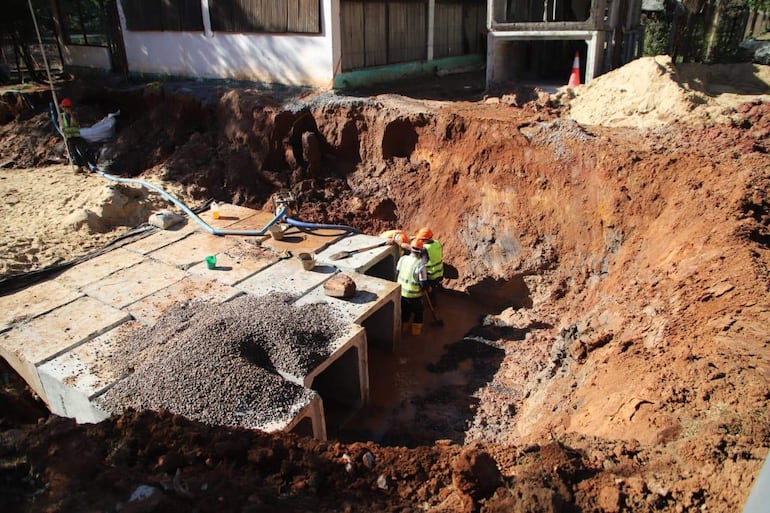 Tres trabajadores con cascos y chalecos reflectantes operan cerca de un área excavada, rodeados de escombros y un tubo azul de drenaje.