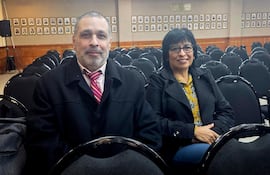 Liliana González de Bristot, con chaqueta negra y blusa amarilla, y Mario Elizeche, con abrigo negro y corbata de rayas, conversan en un auditorio vacío.